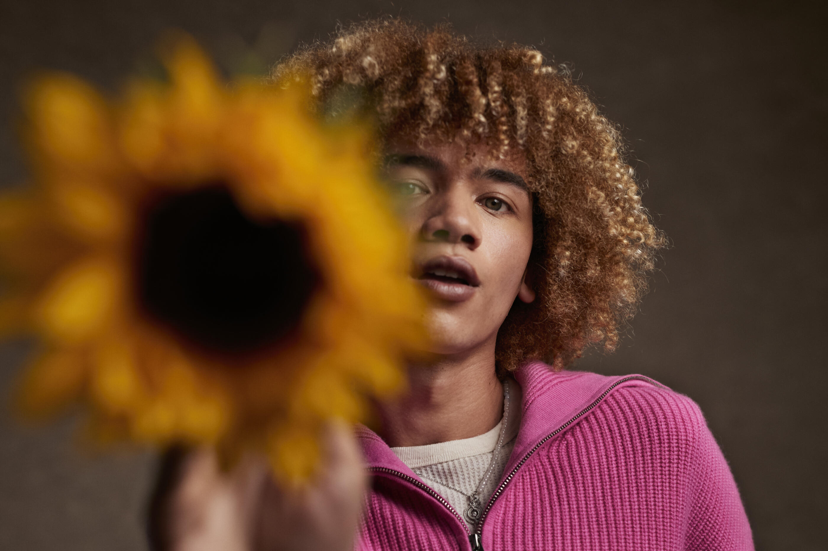 Portrait of curly haired androgynous person showing sunflower against brown background
