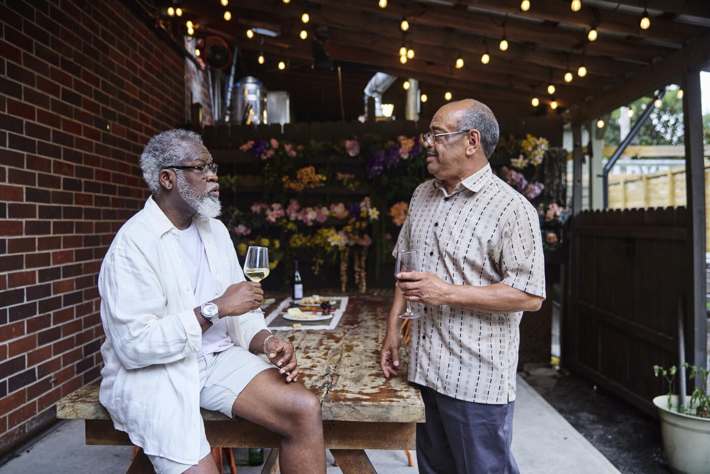 Mature man drinking wine while talking with male friend standing near table in restaurant. They are in a casual setting on covered patio at a local wine bar.