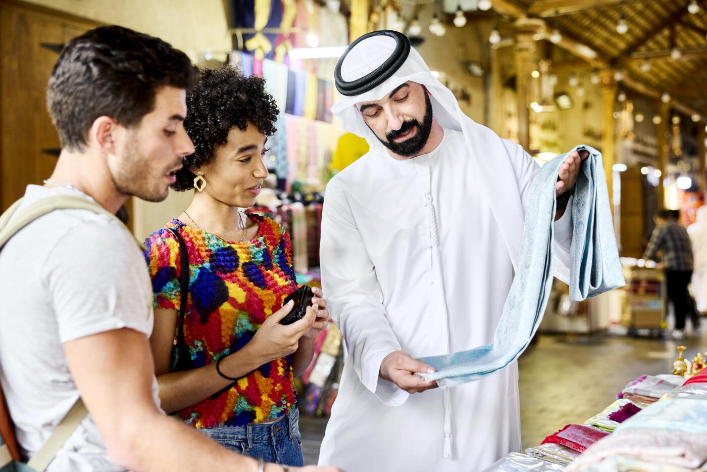Muslim tour guide showing premium fabric to travelers on vacation. Tourists are exploring dye market. Woman is holding camera in her hand.