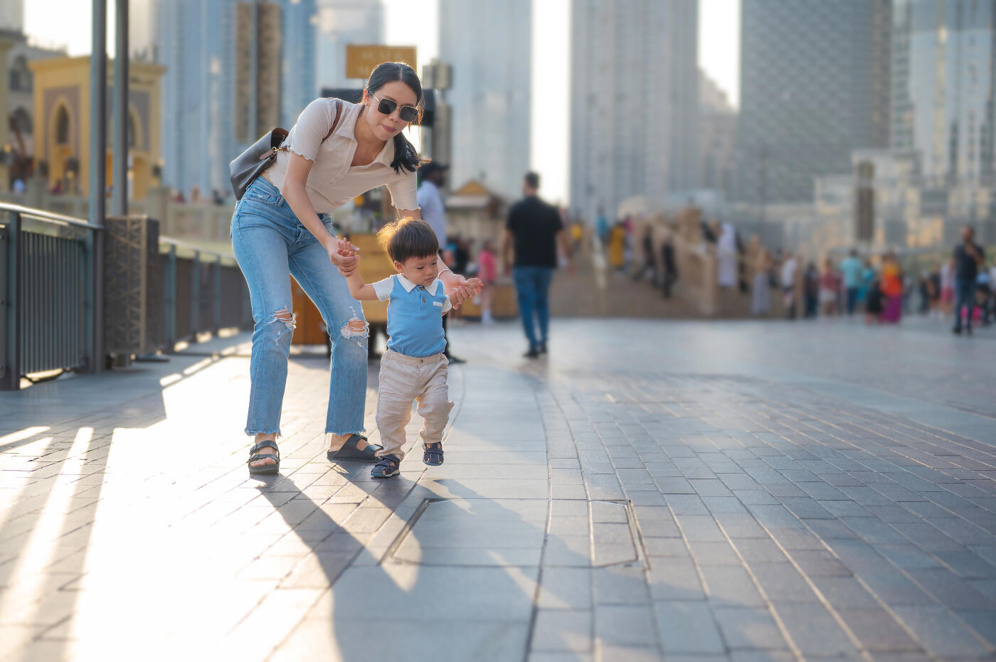 A loving mother assists her young son as he takes steps, exuding joy and excitement in a picturesque urban locale, representing the cherished moments in childhood and the guidance provided by parents.