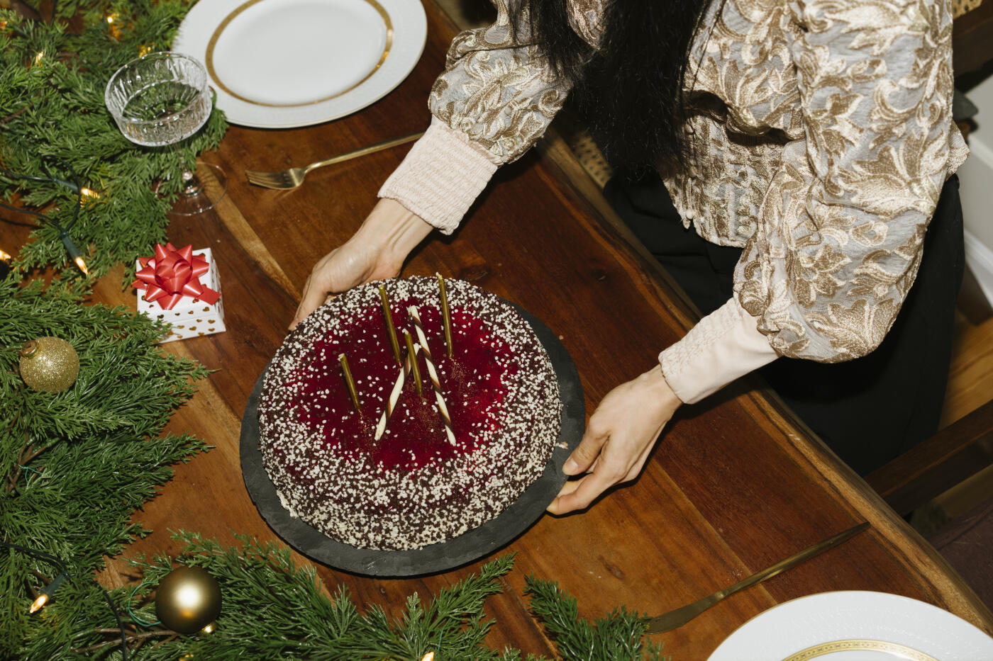View of two arms placing a cake on a table to the left of Christmas tree baubles and fir branches