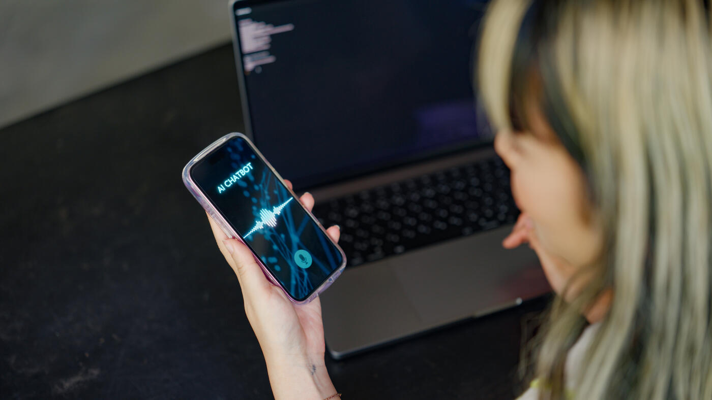 A businesswoman is stuck at work on her laptop. She asks her smartphone's chatbot app for advice and gets a solution, so she proceeds with her work.