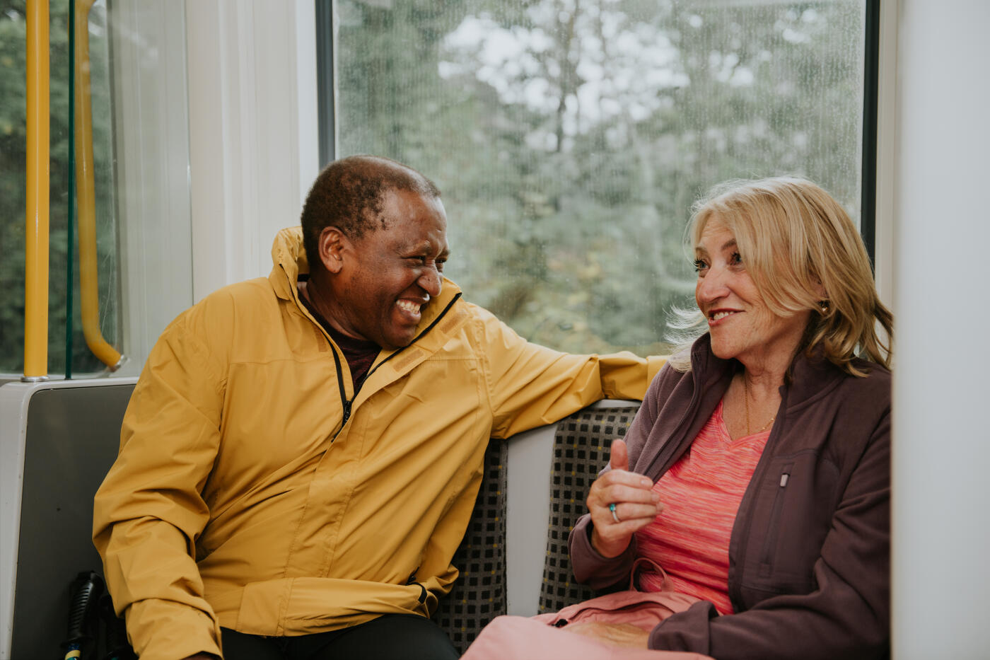 A mature couple in casual outdoor clothing sit beside one another on a train, chatting as they travel to their destination.