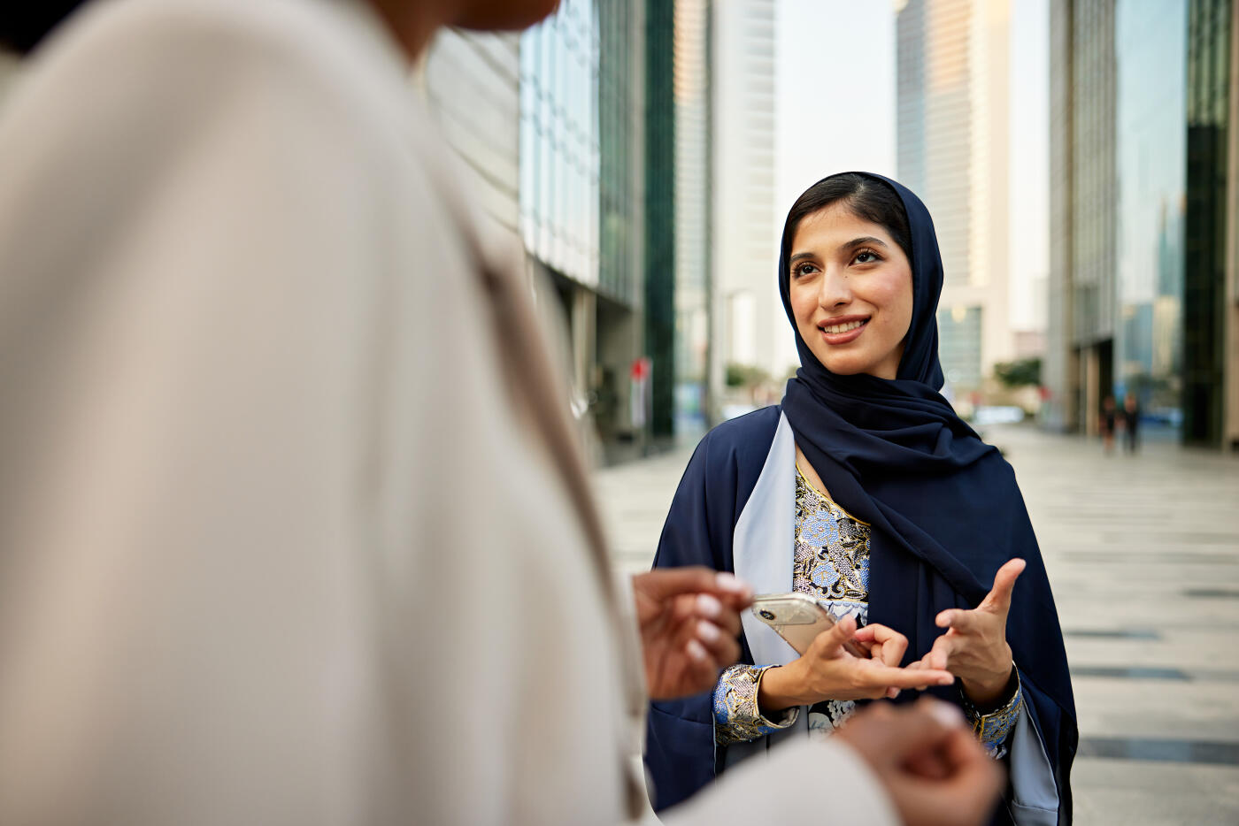 Focus on young woman in traditional attire standing in modern plaza with colleague, smiling, gesturing, making a point.