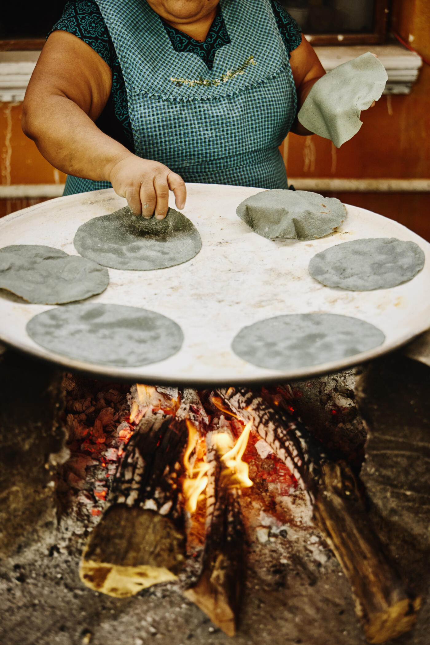 Medium shot of Mexican woman placing traditional blue corn tortillas on wood-fired comal grill to heat them