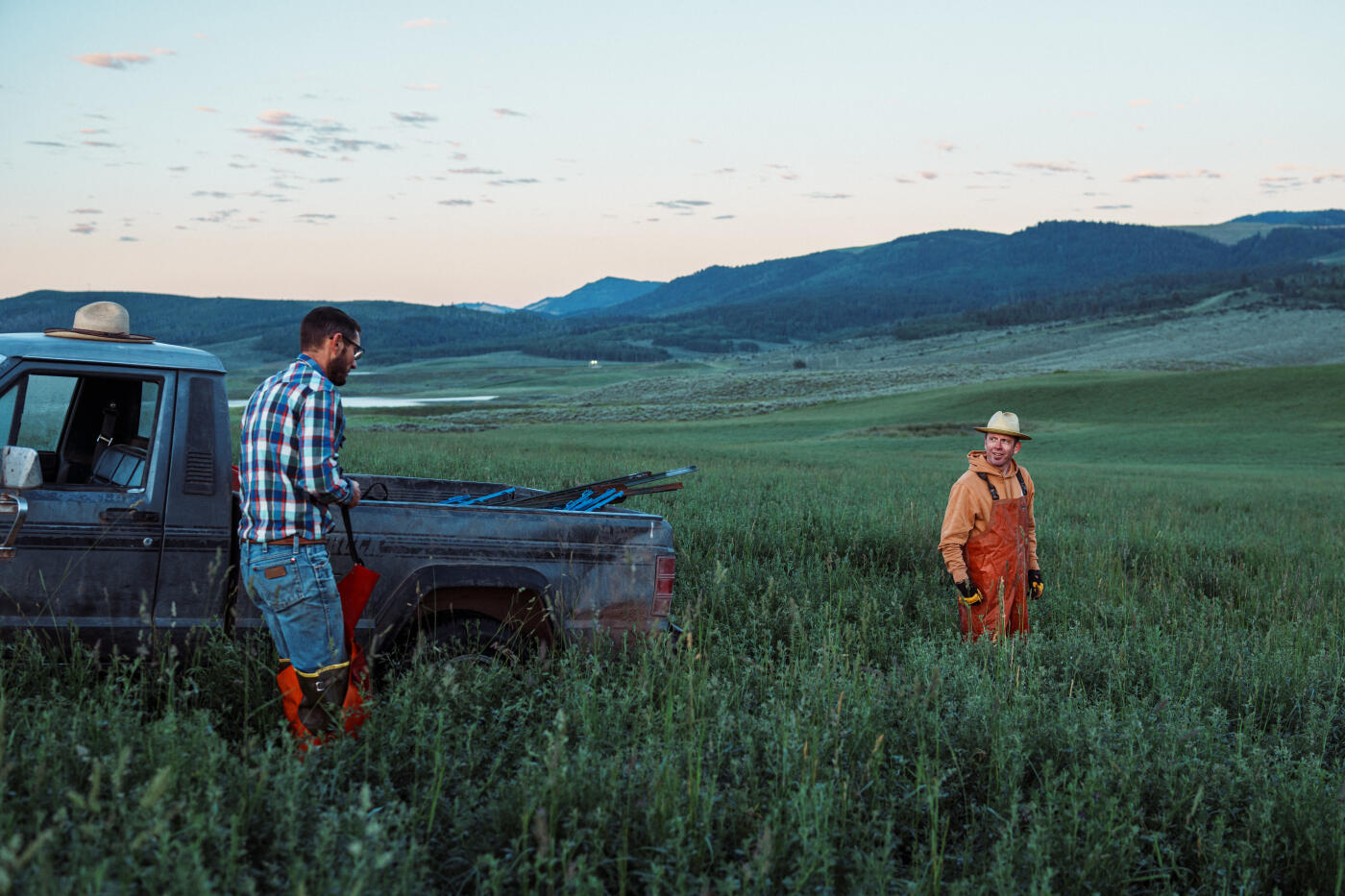 At sunrise, two ranchers unload fencing materials from a pickup truck in a dew-covered hay field nestled among Wyoming’s mountains.