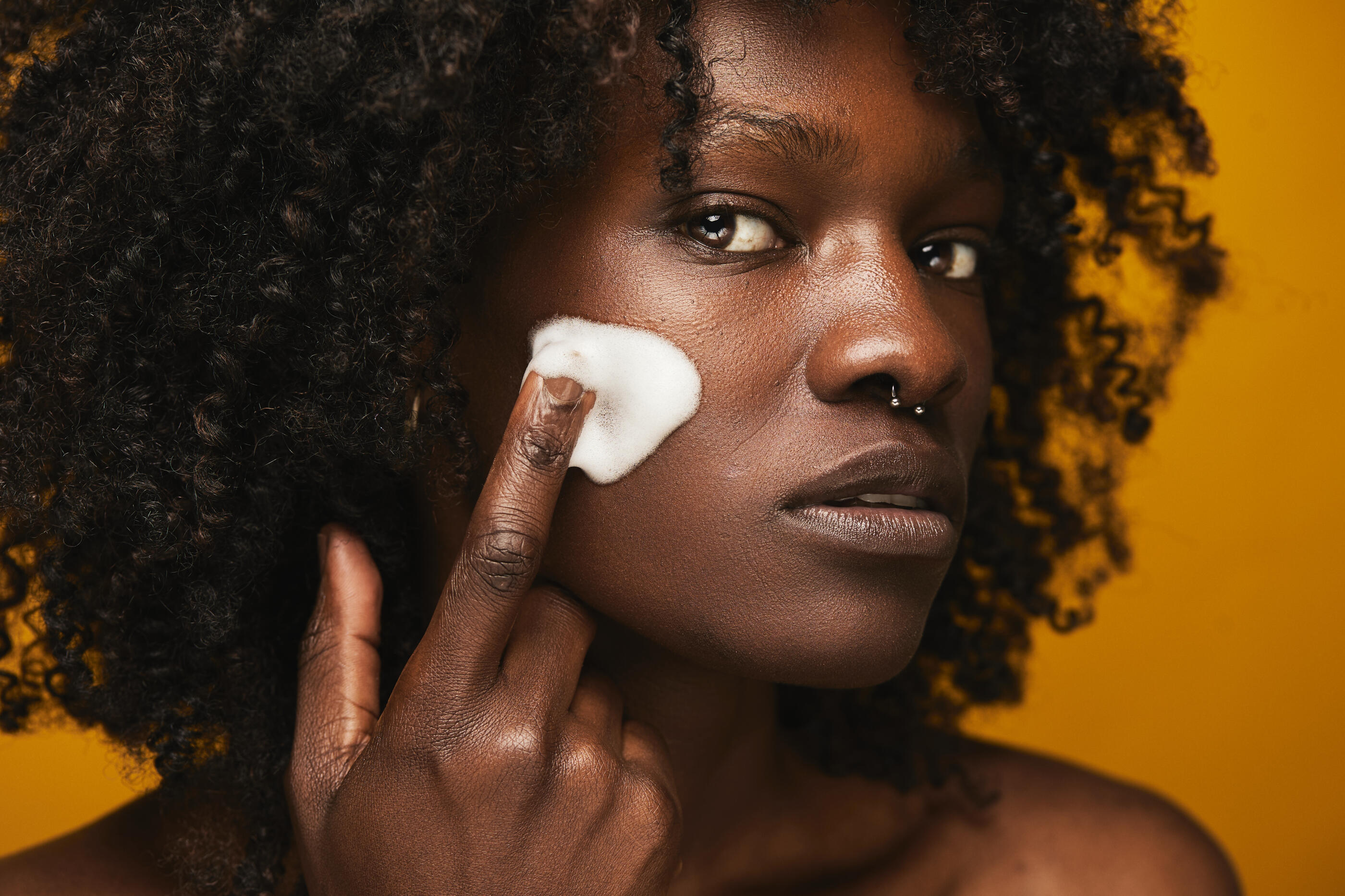 Young adult woman with dark skin and curly hair applying a foamy cleanser to her cheek, focusing on a skincare routine for a clean and fresh complexion against a yellow background