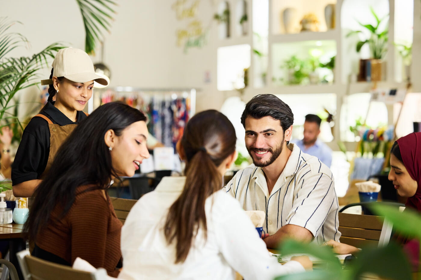 Smiling man with friends at social gathering in coffee shop. Young male is looking at females discussing menu while sitting at table. Waitress is waiting patiently for taking order.