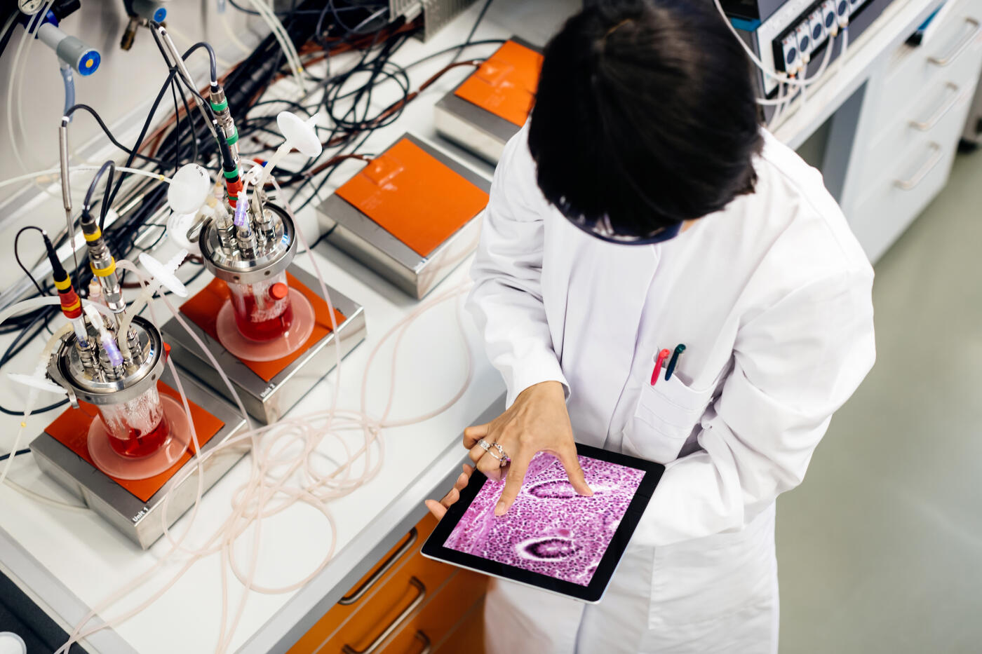 A female asian scientist is doing research in a laboratory. She's working on a tablet computer next to a desk with flasks filled with substances and other scientific equipment.