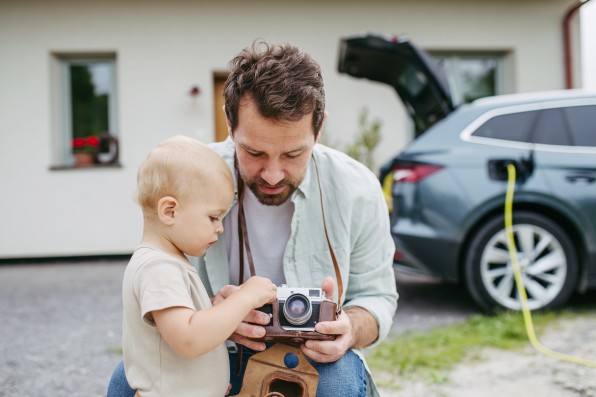 Father showing his old camera to little son. Electric car with charger behind them. Photographer's legacy.