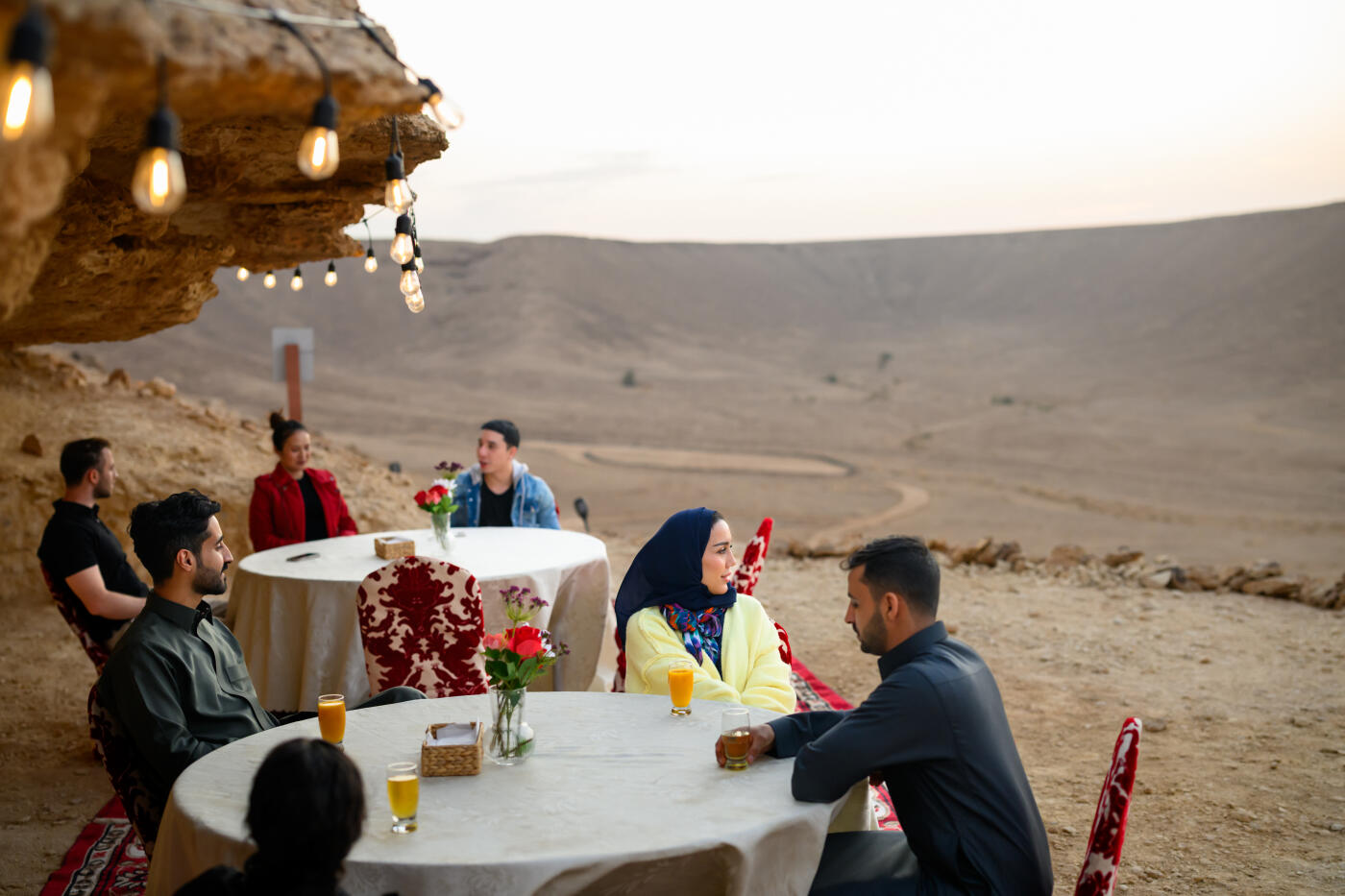 Group of tourists seated at clothed tables beneath a desert cave with hanging lanterns, sharing dinner with expansive sandy landscape visible behind them. Warm golden light illuminates the gathering as evening settles over the terrain.