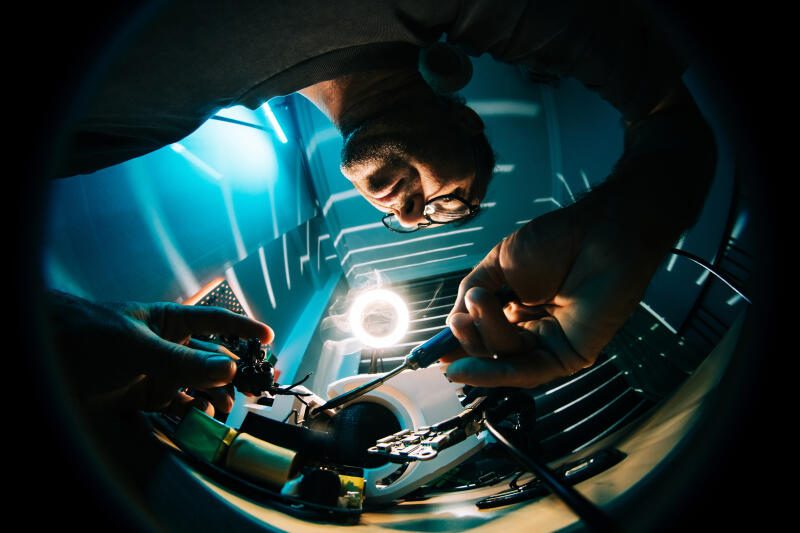 An engineer solders electronic components onto a circuit board, viewed from an intense low-angle perspective.