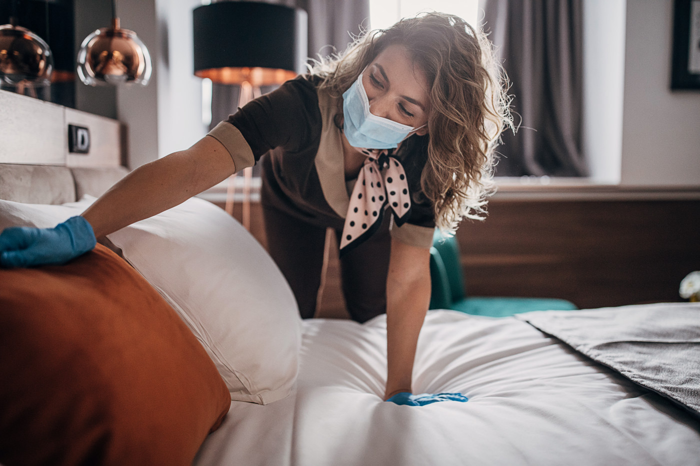 Beautiful maid with medical mask changing bed linen on the bed in a hotel room