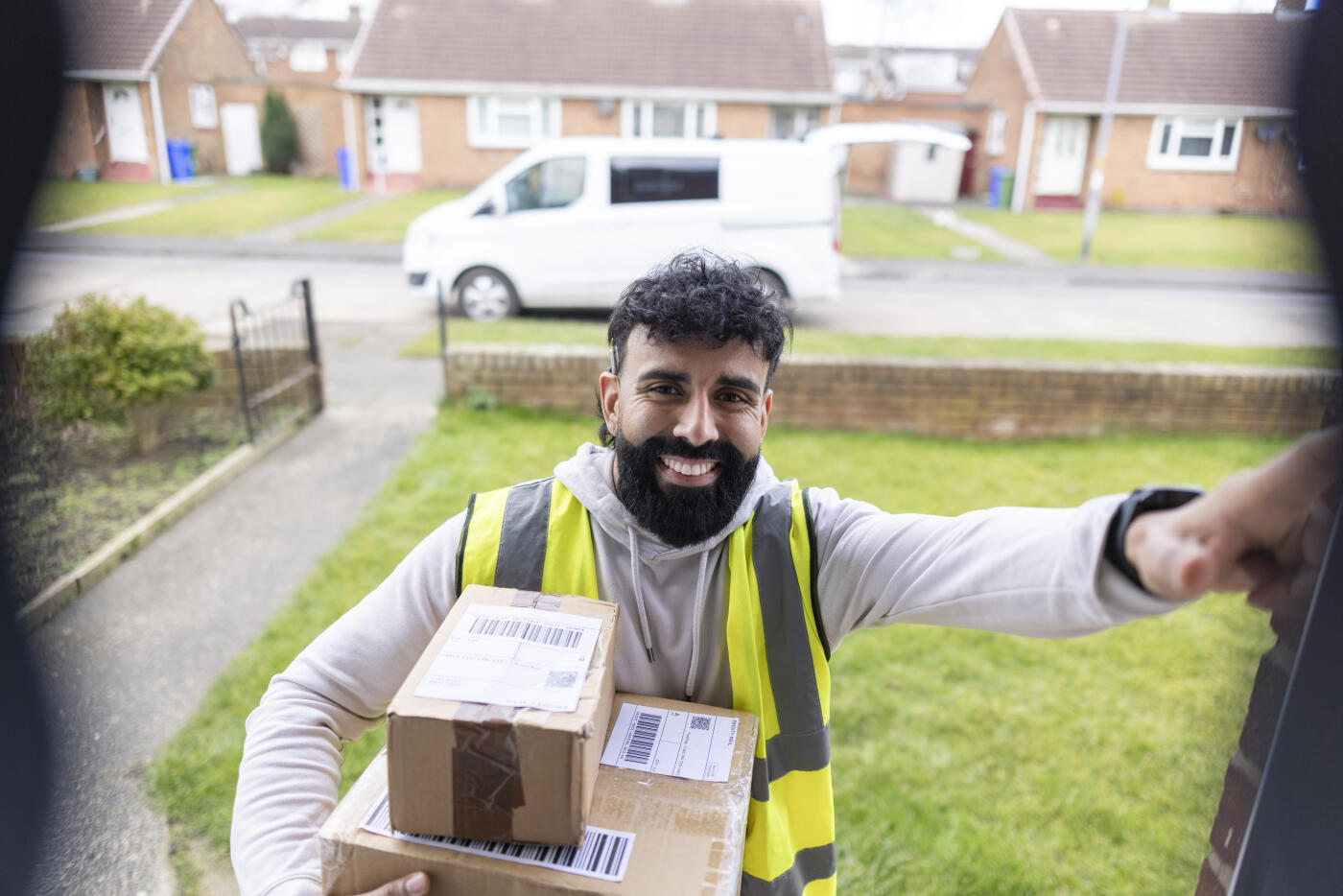 A friendly delivery driver stands at the doorstep, holding a package while looking directly into a smart doorbell camera. Captured from the perspective of the doorbell, the image highlights modern home security and contactless delivery services.Videos are available similar to this scenario.