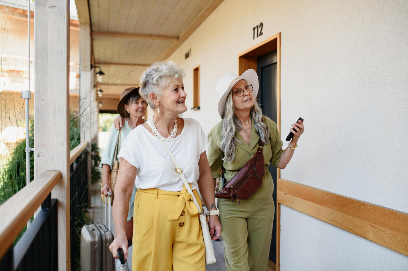 Group of senior female friends with luggage holding smartphones outdoors at hotel terrace.