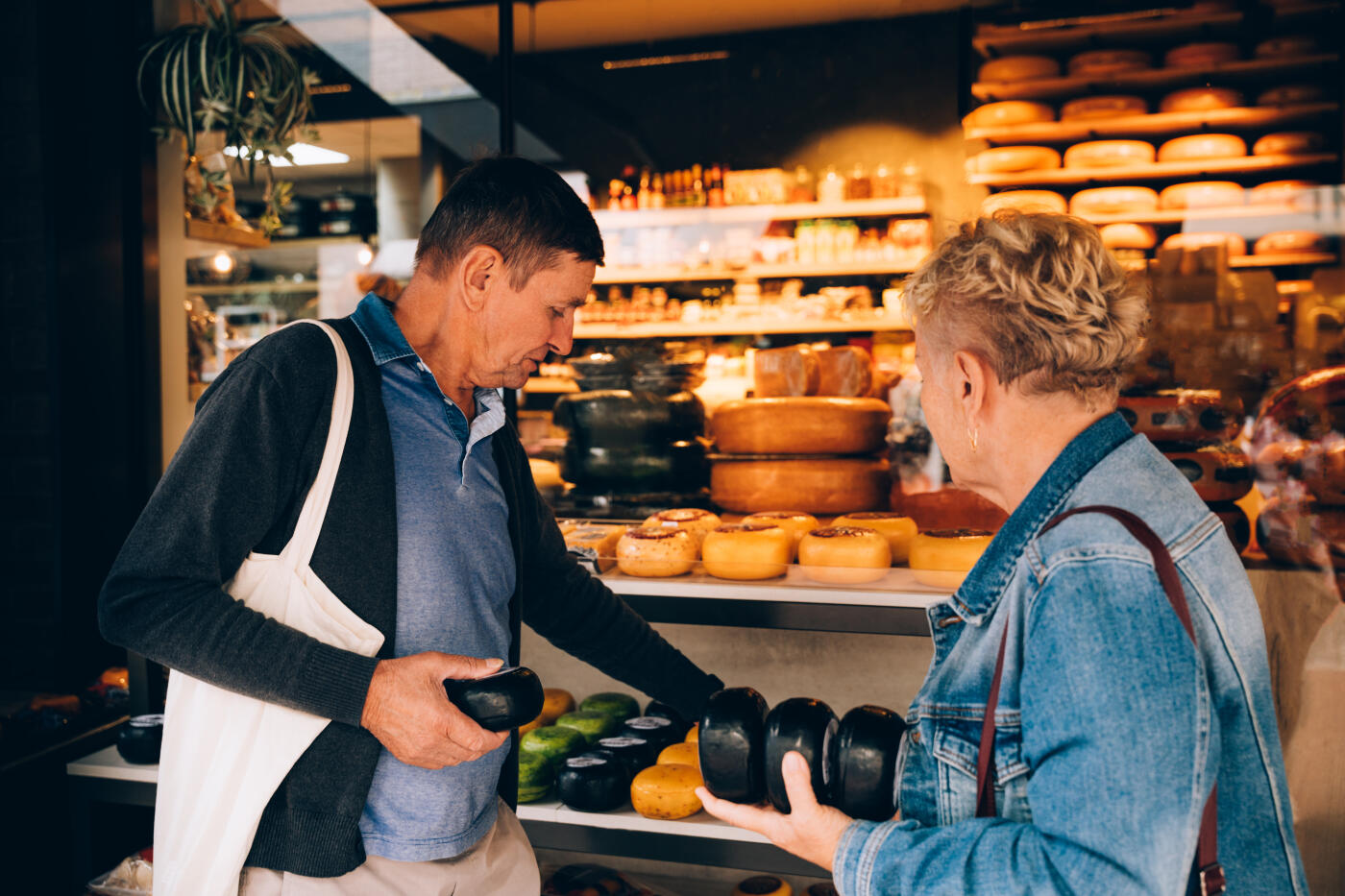 An older couple shopping for cheese at a local store, carefully examining traditional wheels of cheese in various colors and sizes. The display is filled with artisanal cheese varieties, creating a warm and authentic atmosphere that reflects food culture, local traditions, and slow travel experiences