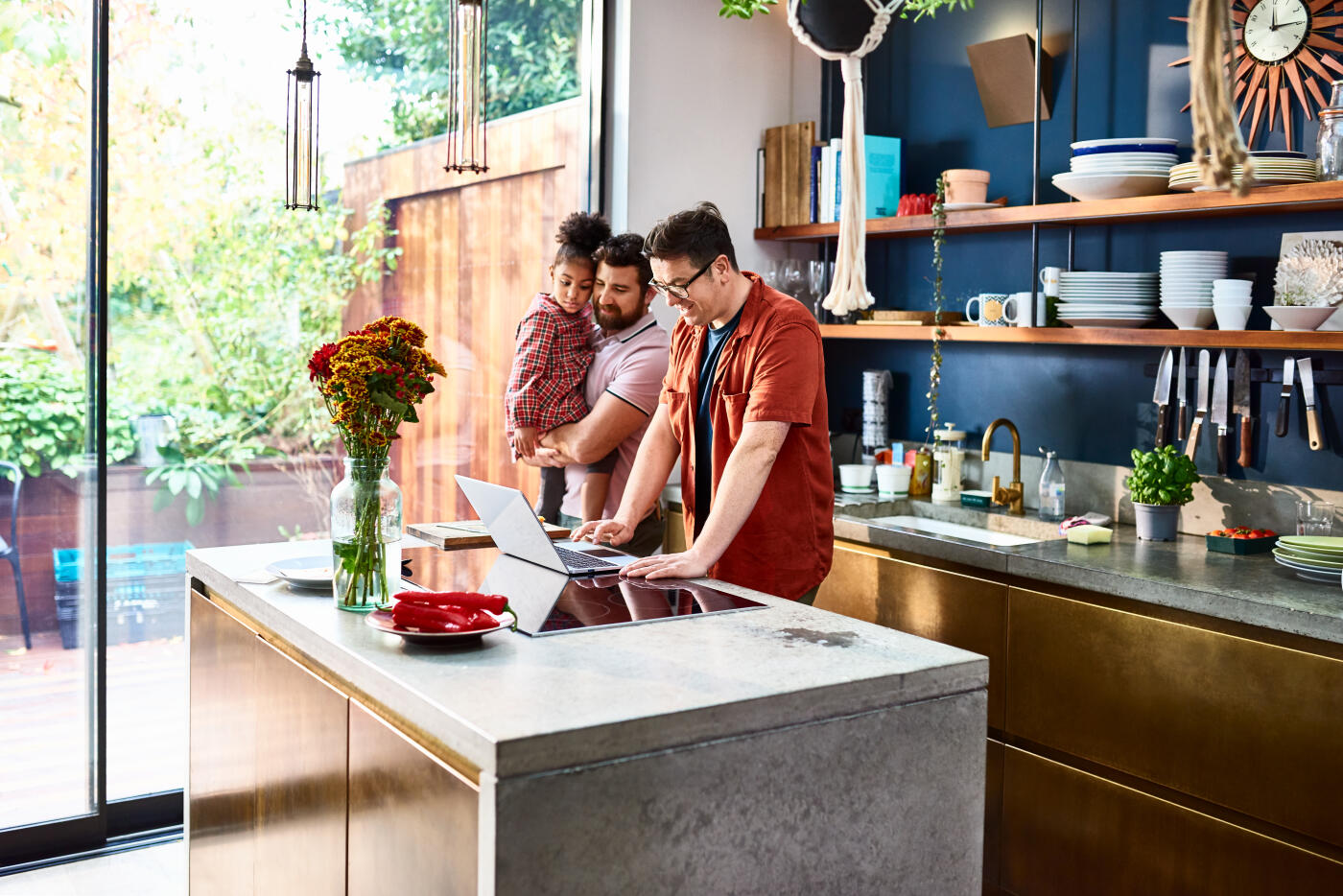 Man holding young daughter at home in kitchen watching partner use laptop, family connections, lockdown, working from home