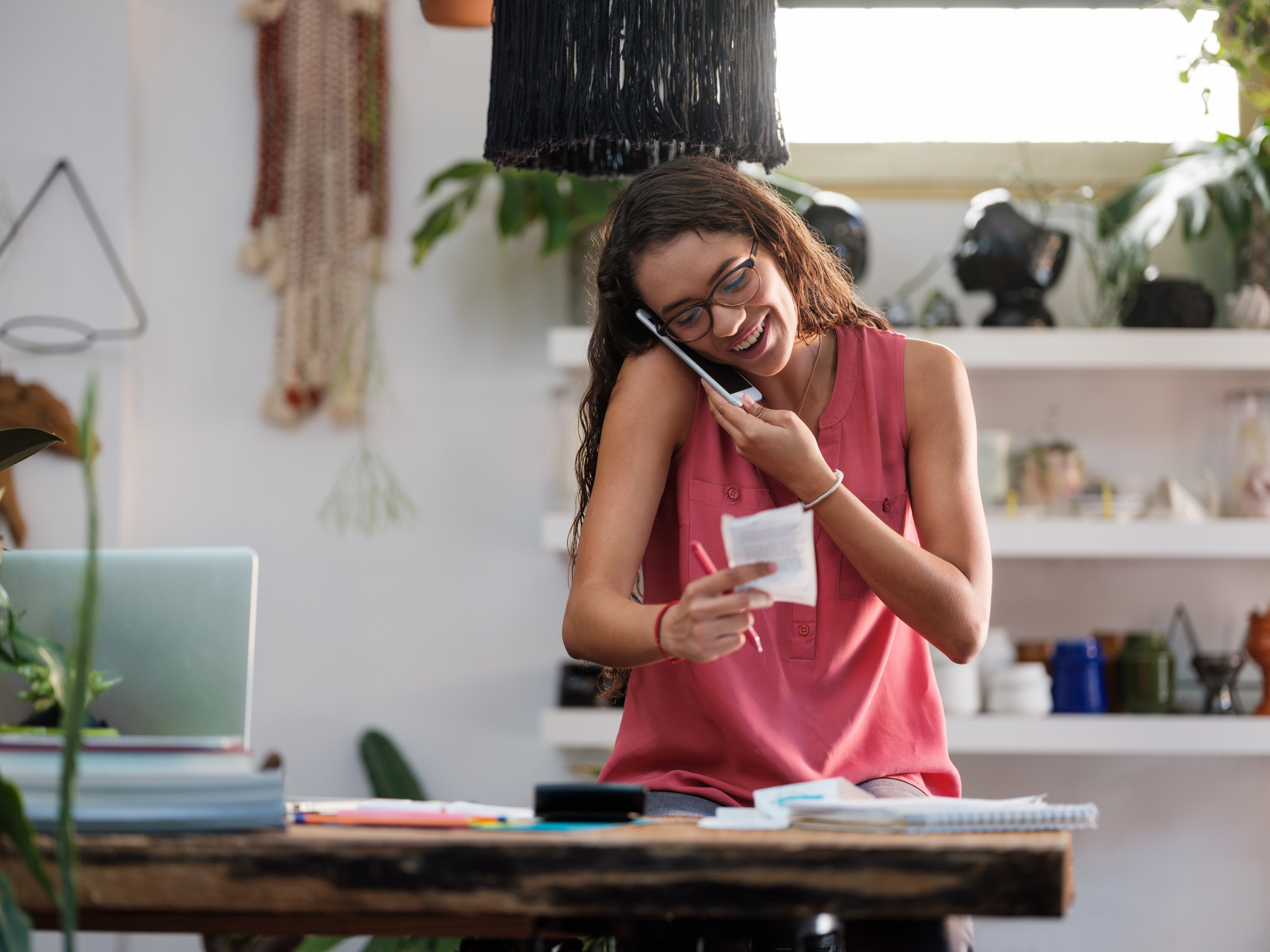 A young female sitting at a plant store holding the phone with the opposite to her ear and looking at a store receipt while smiling.