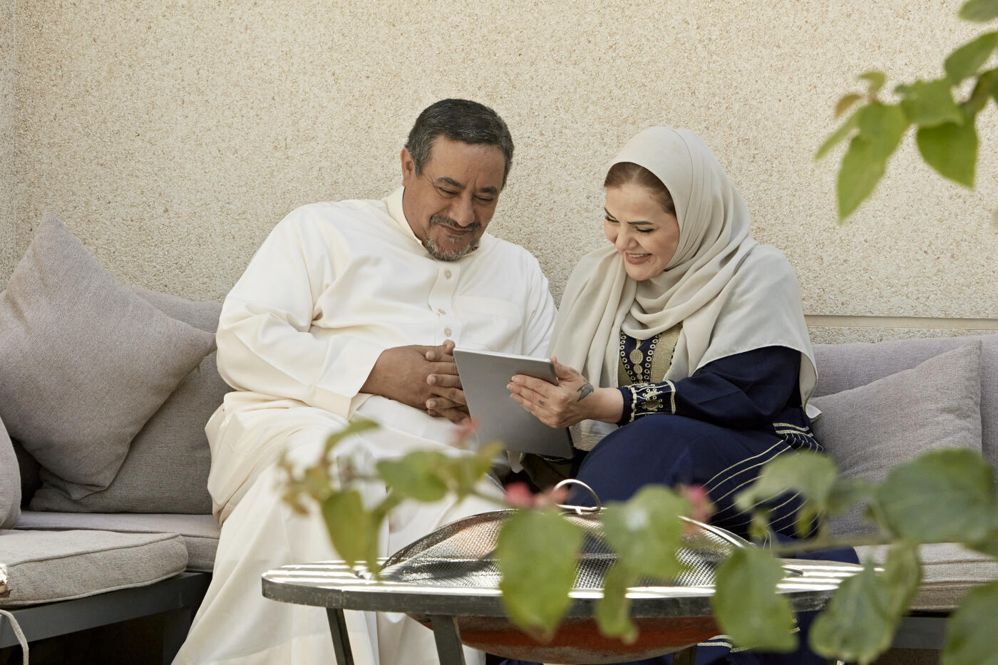Front view of Middle Eastern man and woman in traditional attire framed by foliage and smiling as they sit together on patio using digital tablet.