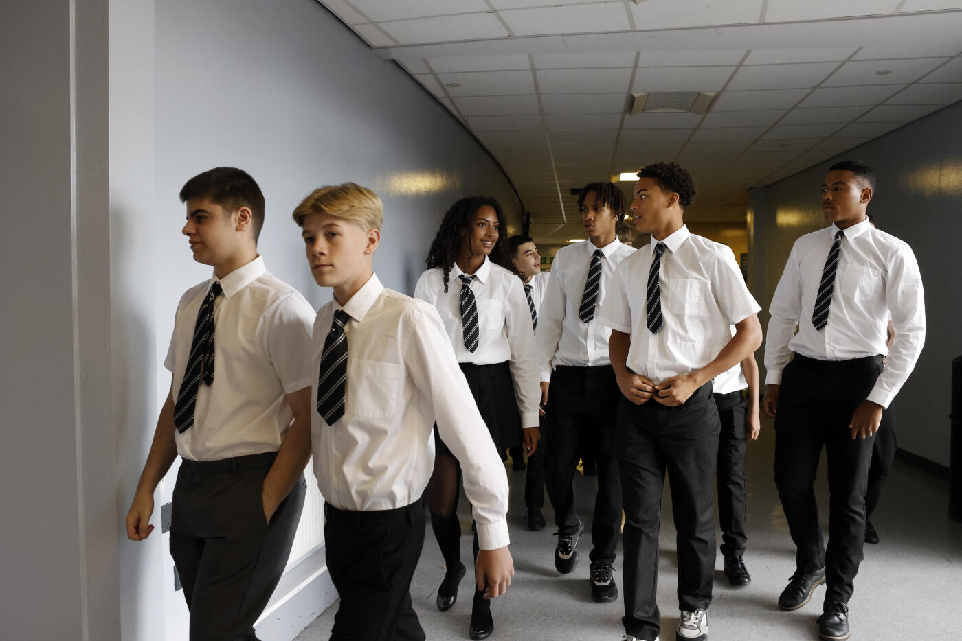 A mixed group of secondary school students in uniform walks out of an examination hall, moving together toward the exit. Their expressions show a mix of relief and quiet reflection as they leave the hall behind.