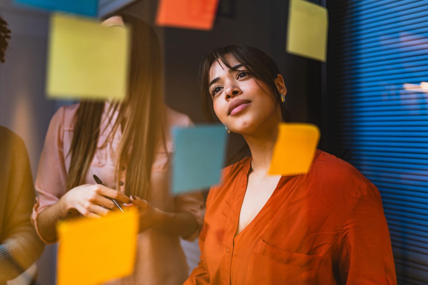 Businesswoman in an orange shirt thoughtfully reading notes on a glass wall during a brainstorming session, highlighting collaboration and strategic thinking in a modern office