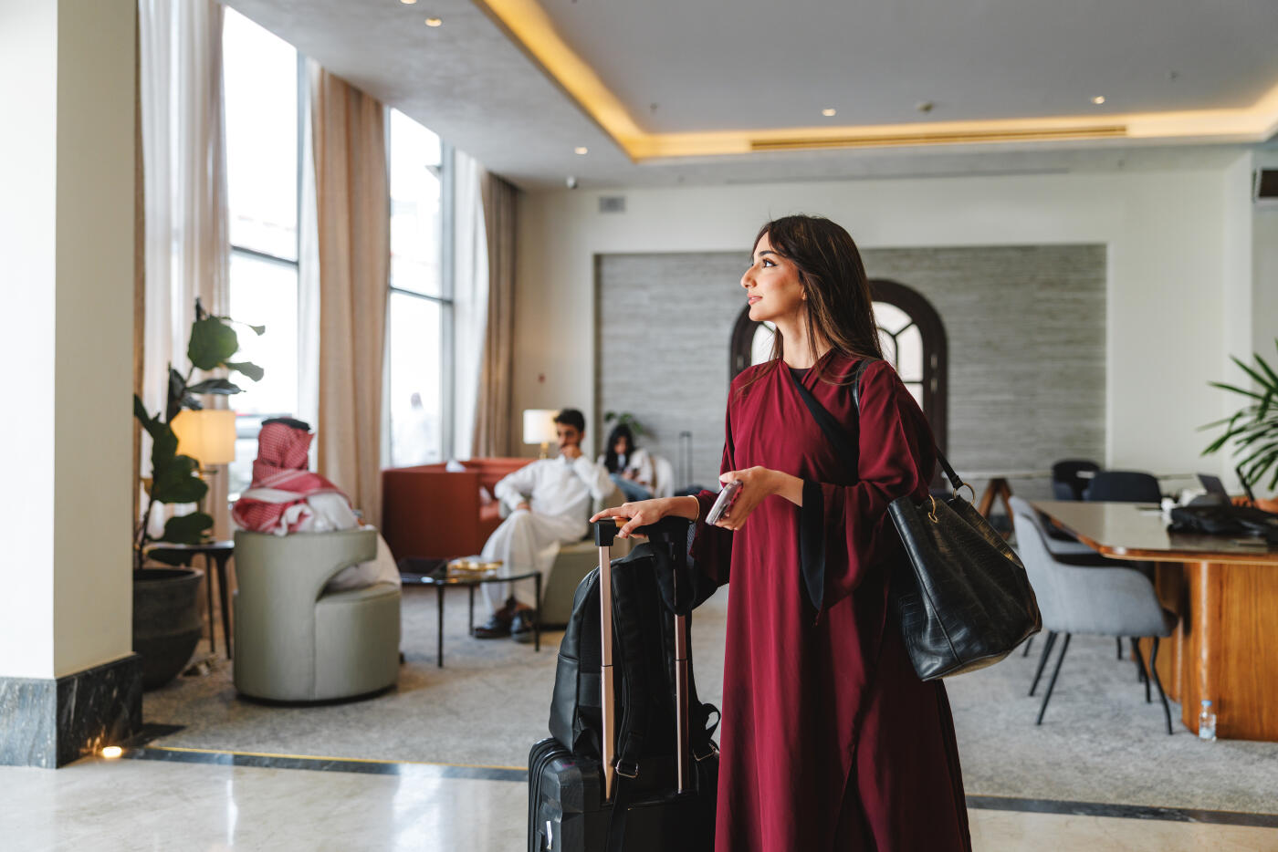 Mid adult Middle Eastern female in an abaya confidently using a smartphone in a hotel lobby. She stands beside her luggage, surrounded by elegant furnishings and soft lighting, conveying a sense of relaxation and purpose.