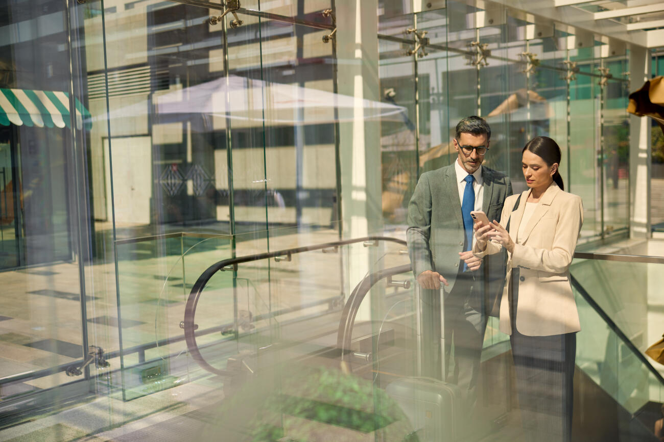 Two individuals in suits are inside a modern glass lobby, ascending the escalator.