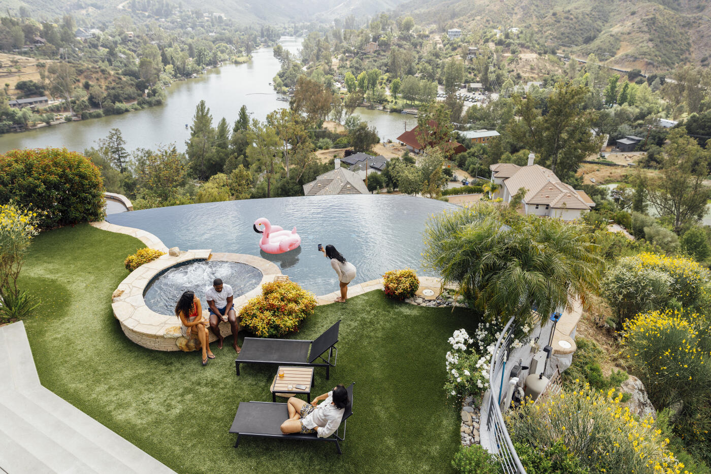 High angle view of a group of multiracial male and female friends spending leisure time in a backyard near a swimming pool, captured against a scenic view of lush trees and natural surroundings. The group is relaxing outdoors during a summer weekend gathering. They are engaged in conversation and enjoying refreshments near the water, emphasizing themes of friendship, luxury lifestyle, and togetherness.