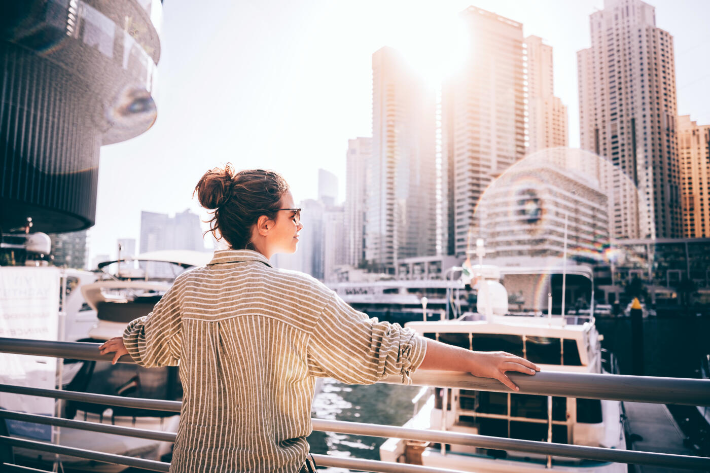Stylish young woman in sunglasses standing by the waterfront in Dubai Marina, enjoying the warm sunlight with a smile. Contemporary city skyline with high-rise buildings and reflections in the canal water. Urban lifestyle and travel freedom concept in the UAE.