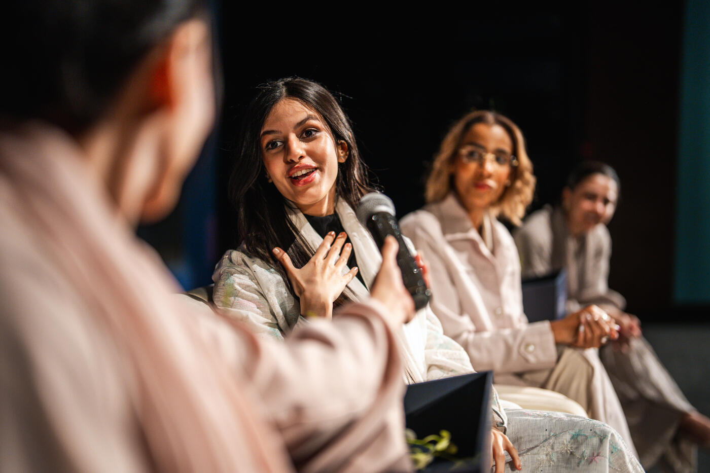 Middle Eastern woman panelist engaging audience while speaking into microphone during inspiring university event in Riyadh auditorium, with attentive listeners in background.
