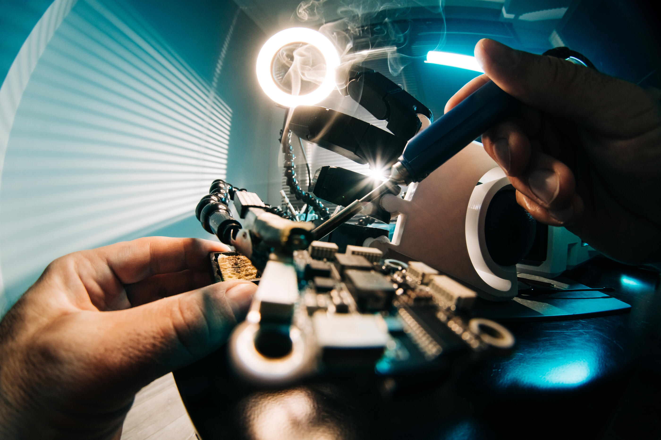 A close-up, wide-angle shot of hands precisely soldering a microchip under a bright ring light with rising smoke. The dramatic turquoise and white lighting conveys the essence of technical innovation, engineering excellence, and high craftsmanship.