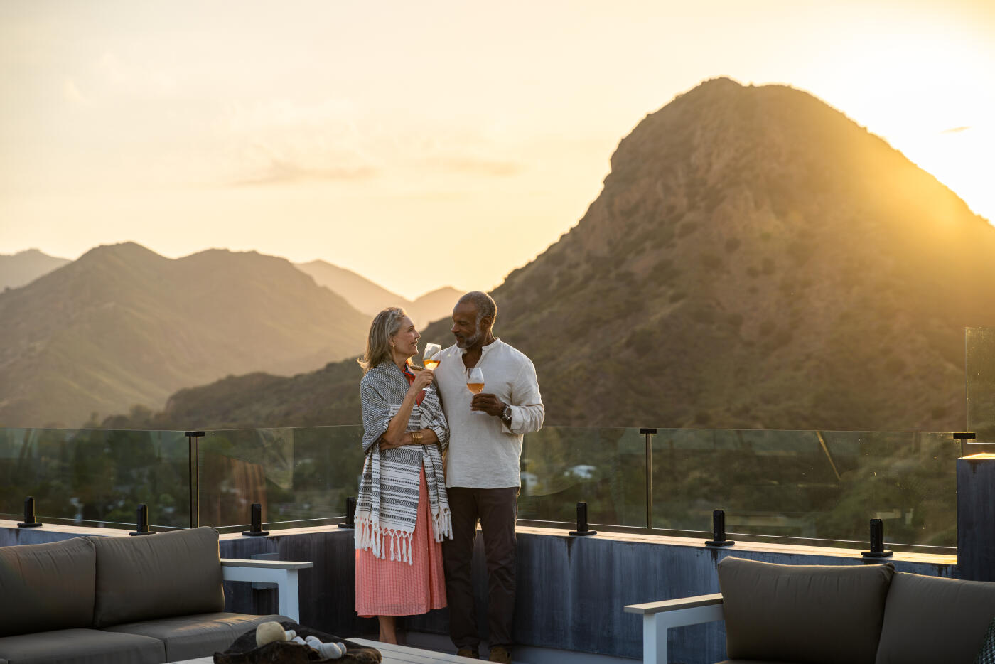 A mature couple stands close together on a rooftop terrace with glass railings, raising glasses of rosé wine in a celebratory toast at sunset. The woman wears a patterned wrap and coral dress, while the man wears a white linen shirt, and both smile as they look at each other. Rolling hills and a prominent peak rise behind them, backlit by warm evening light, with modern outdoor lounge seating in the foreground.
