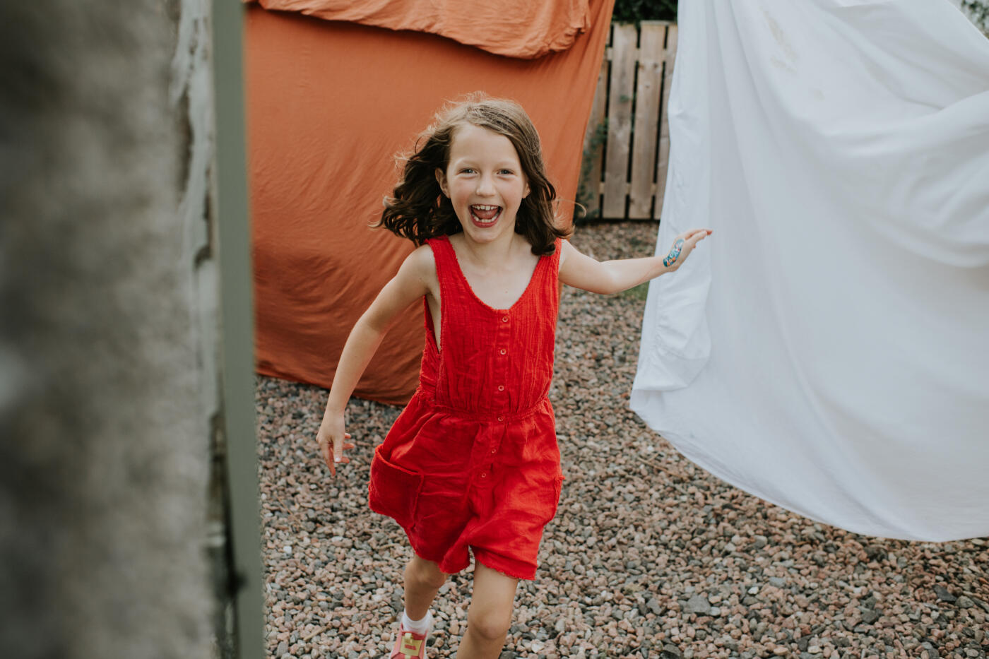 A little girl has fun after freshly laundered bedsheets are hung on a washing line outside. She runs between linen bedsheets whilst laughing.
