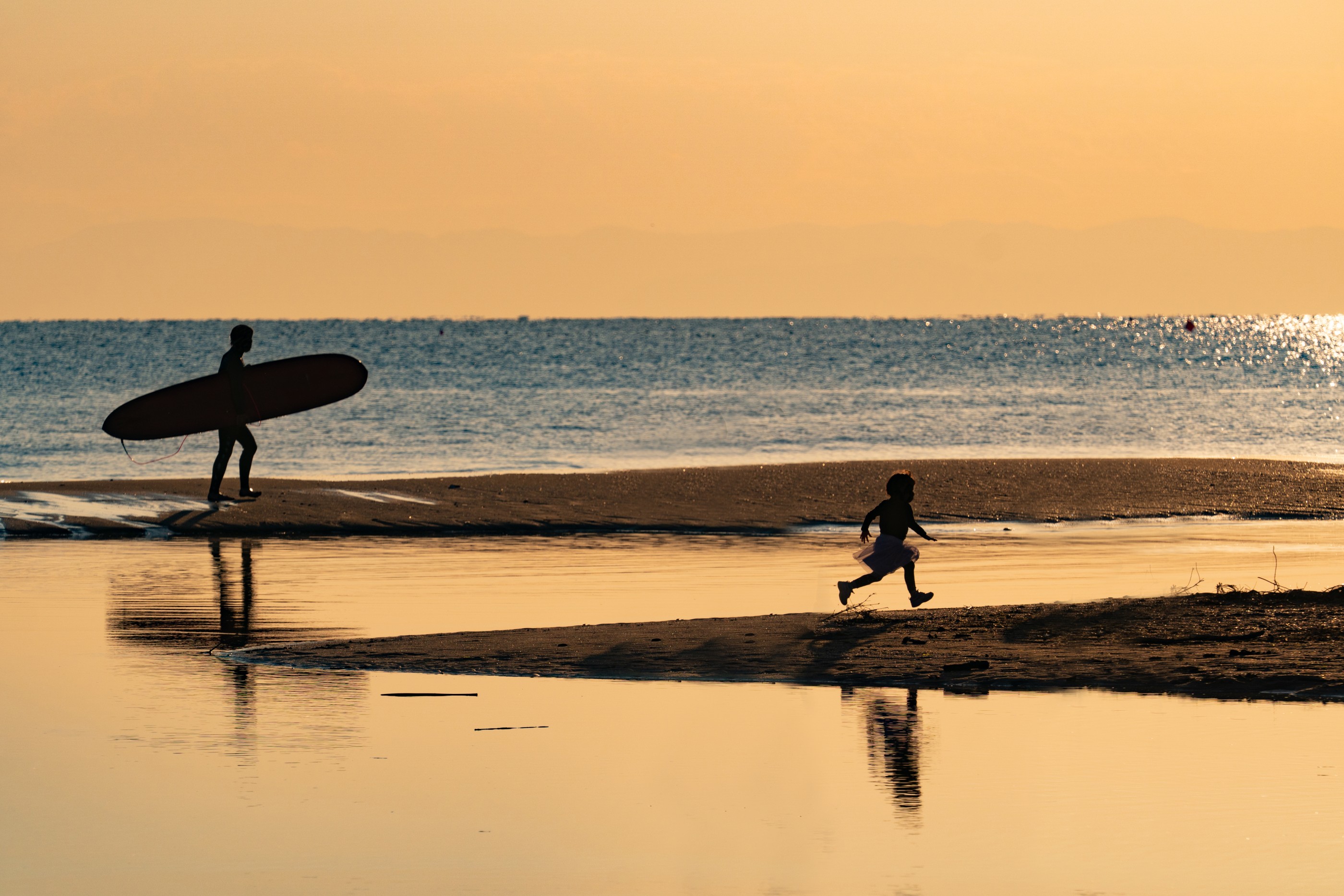 The photo was taken at Yuigahama Beach, Kamakura city of Kanagawa prefecture, Japan. The bottom river is Namerigawa River. The right kid is tourist who are running on the sunset beach and the left surfer is going home after surfing..