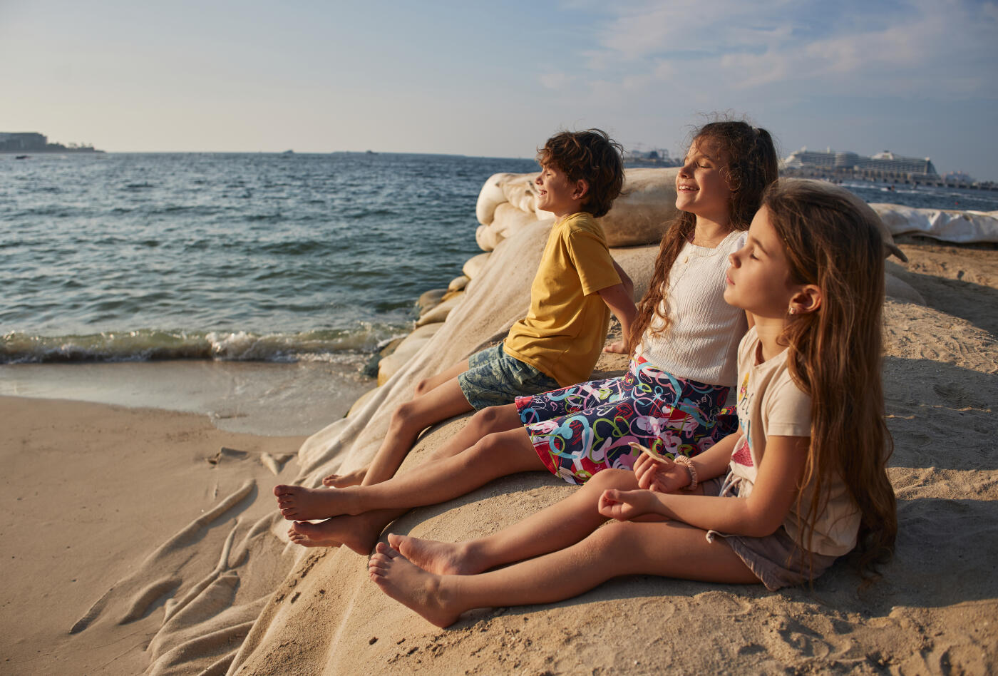 Three young children are sitting on a sandy beach by the sea, enjoying the sun and ocean breeze