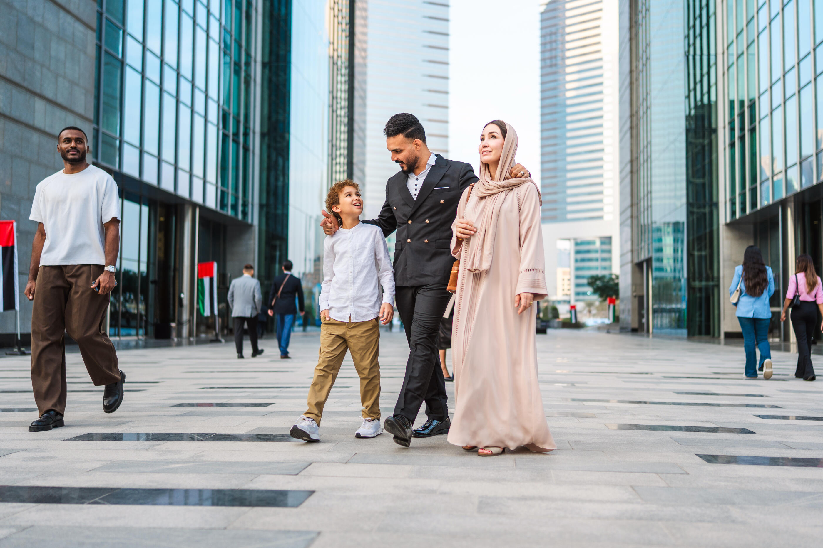 Middle Eastern family strolling through DIFC plaza, with father in business attire, mother in traditional hijab and child between them. Modern glass towers frame the scene under bright daylight, other pedestrians visible in the background.