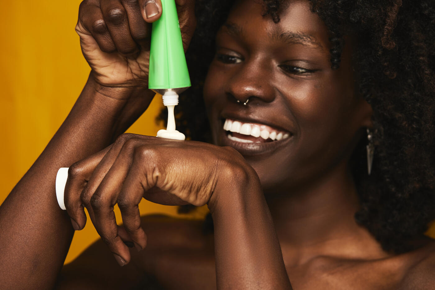 Young adult black woman smiling while squeezing a cream tube onto her hand, focusing on daily skincare and hydrating her skin for a radiant, healthy glow