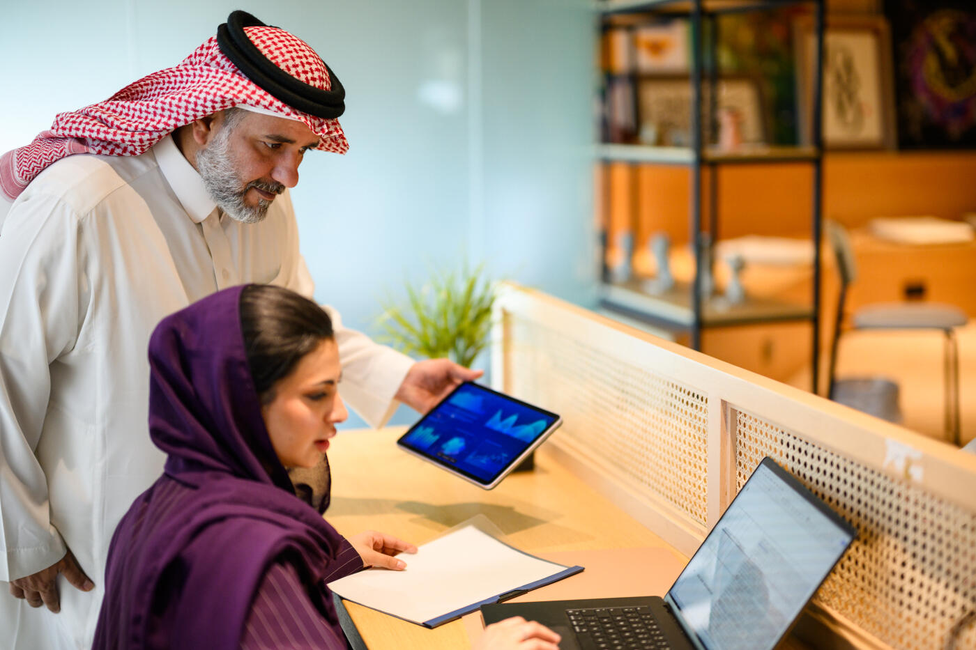 Over the shoulder view of young Arab businesswoman wearing hijab working on laptop while mature male colleague stands beside her in modern office workspace.