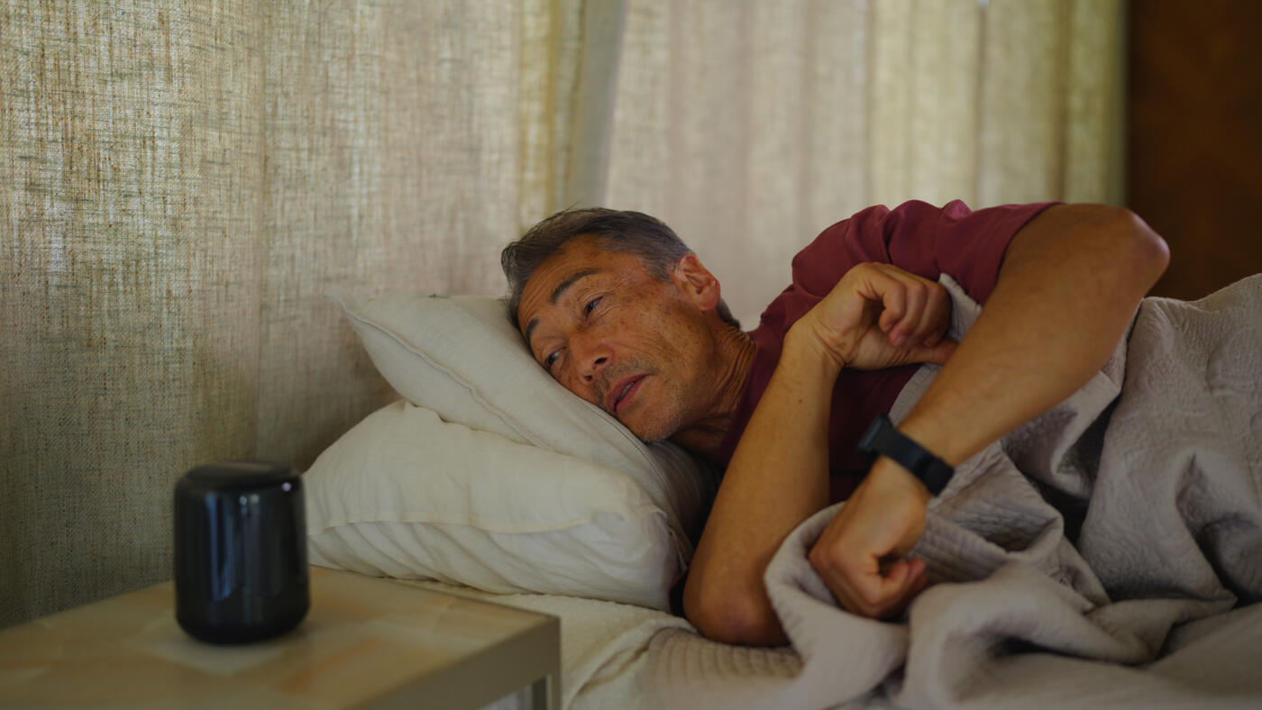 A senior adult man is waking up in his bed and checking his schedule with his smart speaker and smart watch in his bed in the morning in the bedroom.