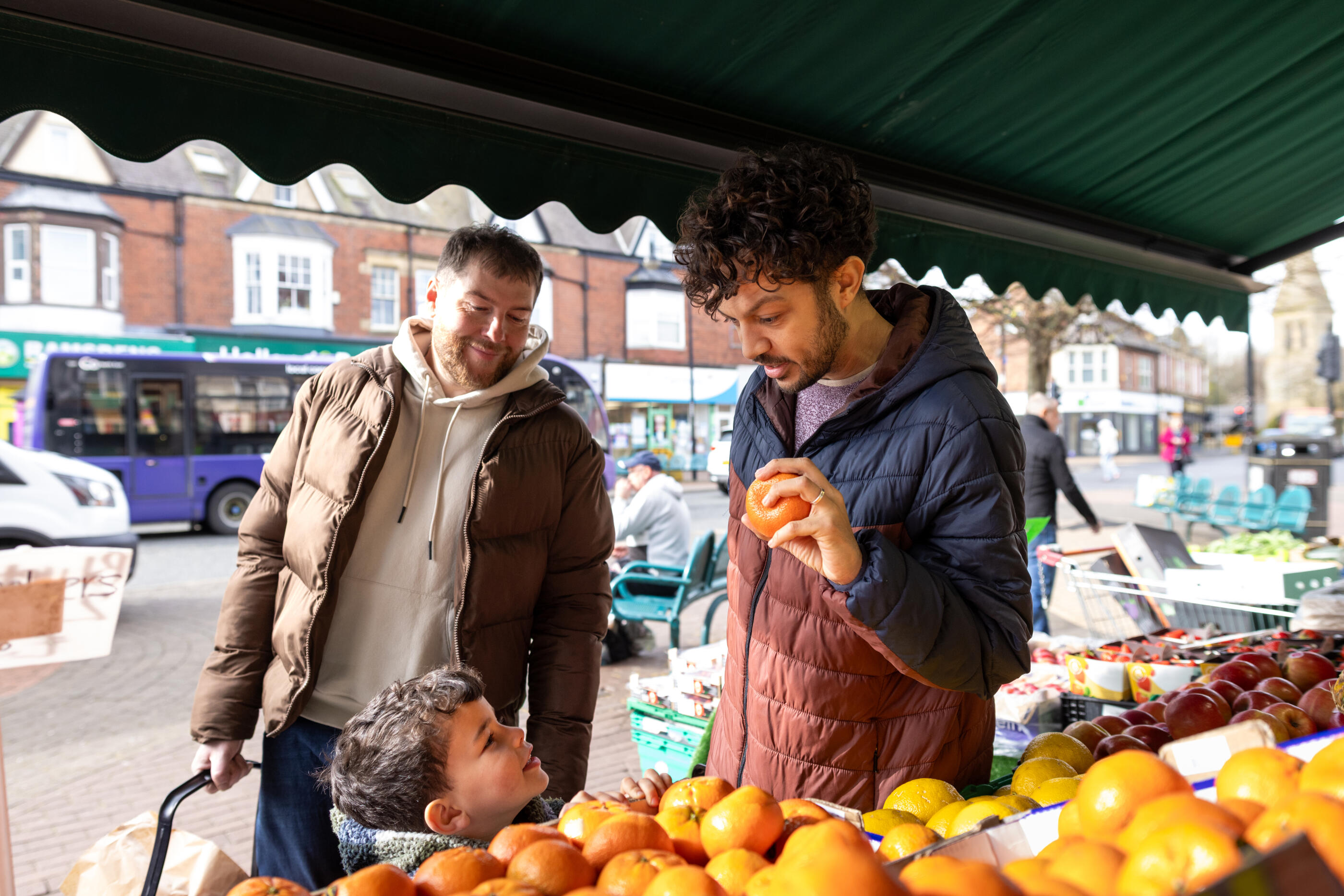 A wide shot of a man standing outside a fruit and veg grocer in Whitley Bay. The man is shopping with his son and partner for healthy ingredients to cook a meal. The son looks up at his father and smiles while the father holds an orange.

Videos are available similar to this scenario.