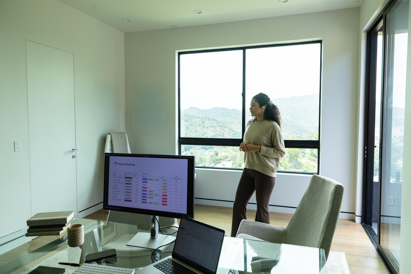 A mid adult woman with curly hair, wearing a beige sweater and wireless earbuds, stands in a bright home office as she talks through a project roadmap displayed on a large monitor. A laptop and glass desk sit in the foreground, while a wall of tall windows reveals rolling green hills in daylight. The minimalist interior with wood flooring and neutral tones suits the hilly landscape of the surrounding area.