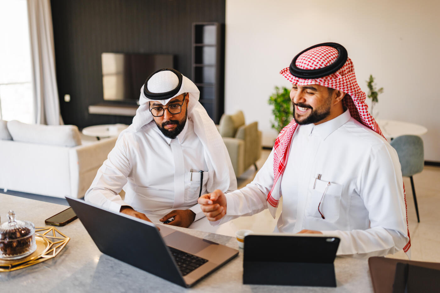 Two Arabic businessmen are collaborating on a project in a luxury hotel room. They are using a laptop and a digital tablet to discuss ideas. The setting is modern and comfortable, promoting productivity and focus