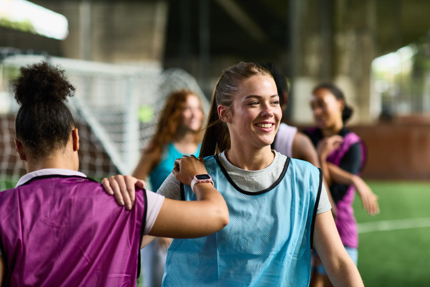Two five a side soccer players in football bibs stretching and warming up before a game, young woman in blue bib smiling and looking away