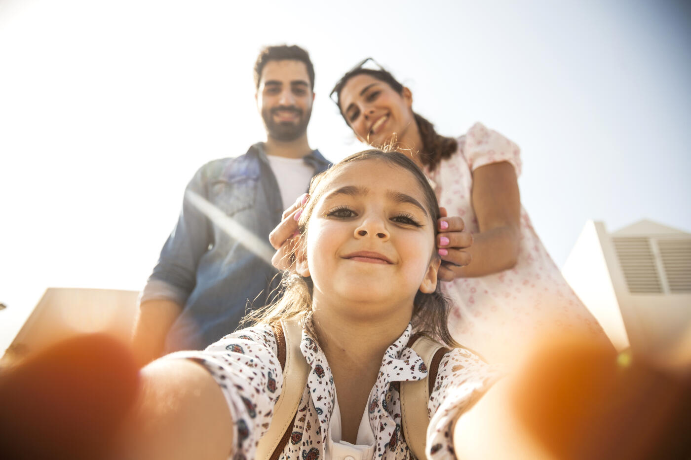 Little girl enjoying the weekend outdoor during a sunny day. She's holding the phone and taking a selfie to her and her parents.