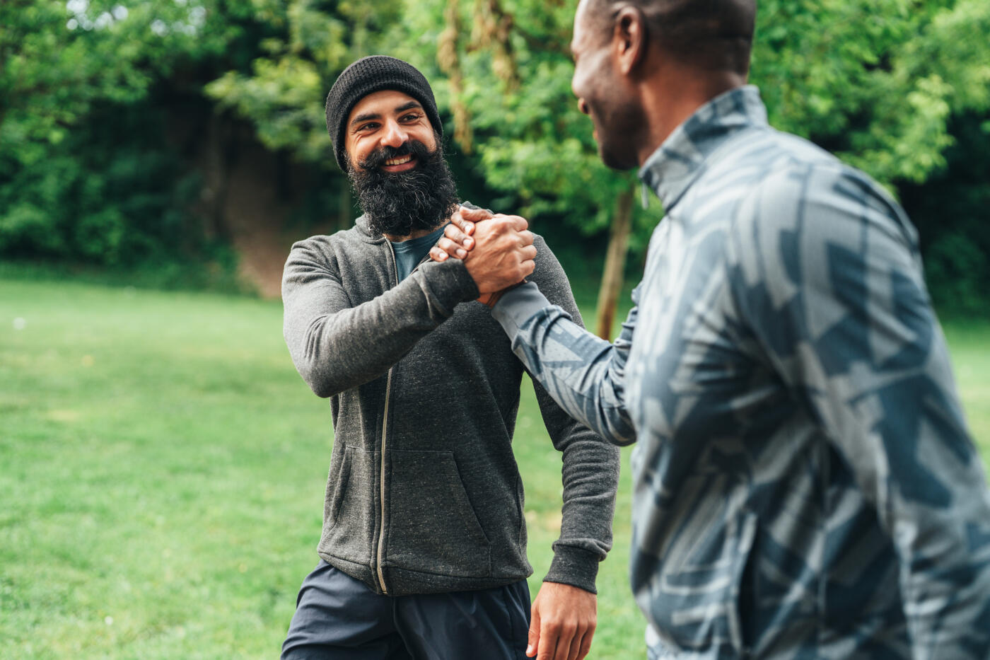 Two male friends share a warm and energetic handshake while smiling at each other in a park setting. Both are dressed in casual athletic wear, suggesting a friendly meetup before or after a workout. The lush green background and natural light create a positive and uplifting atmosphere, emphasizing friendship, connection, and an active lifestyle.