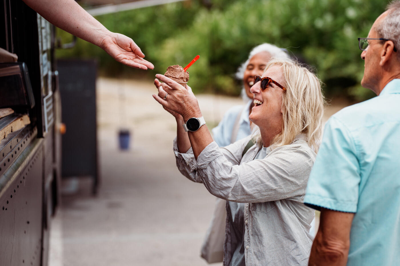 An unrecognizable restaurant employee extends their arm out the window of a food truck and hands a cup of chocolate gelato to a smiling senior woman dining out with her friends.