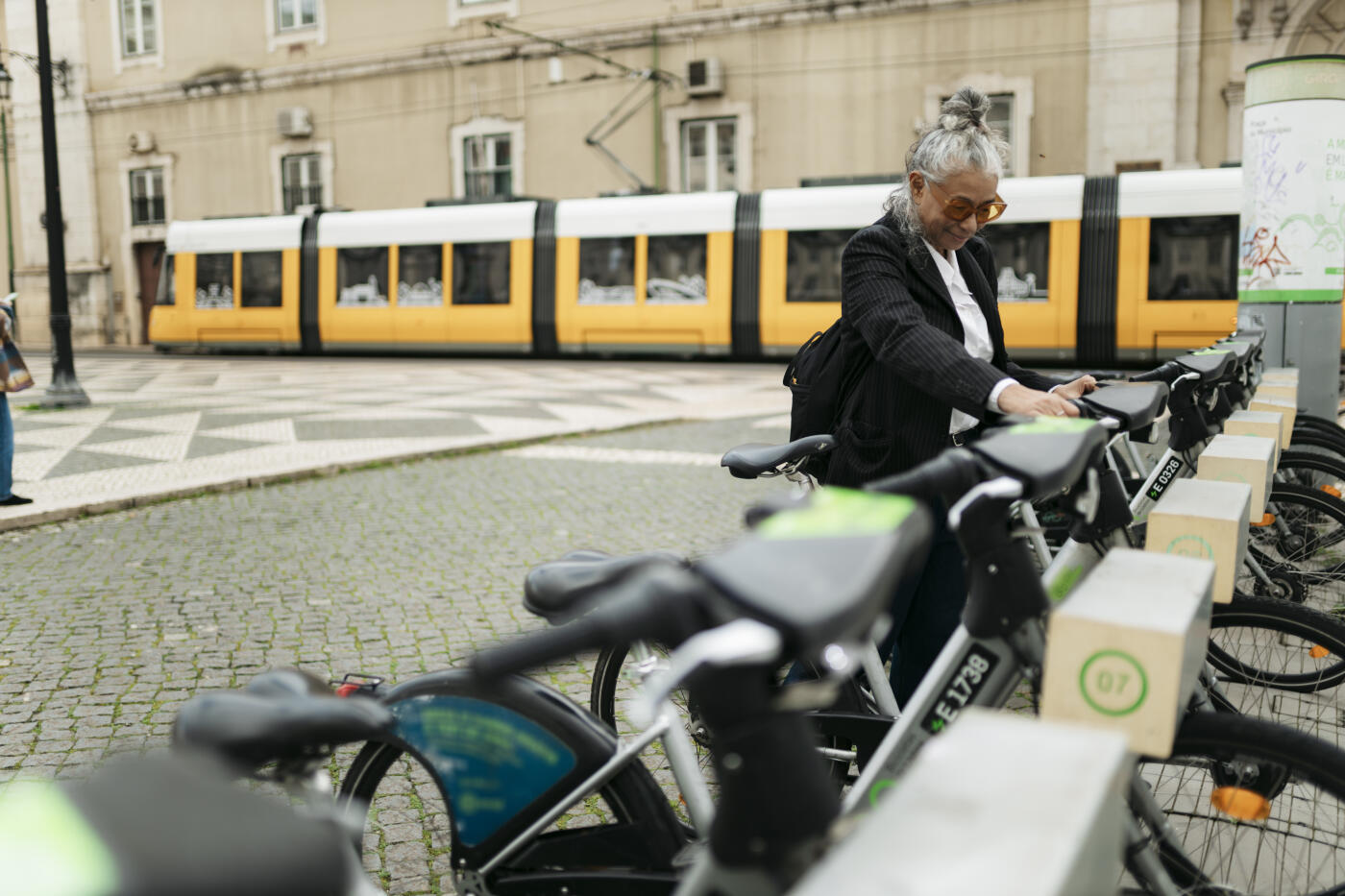 Mature businesswoman renting a bicycle in a bike sharing station in the city, with a tram passing in the background