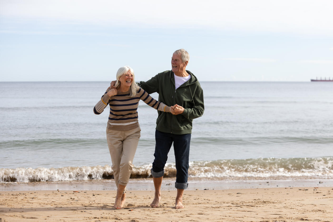 A wide shot of an older couple enjoying a peaceful walk along the shoreline, holding hands and smiling as gentle waves lap at their feet. The serene ocean and sandy beach create a calm, romantic atmosphere, symbolising love, companionship, and the joy of aging together. Perfect for themes of retirement, wellness, relationships, and leisure.Videos are available similar to this scenario.