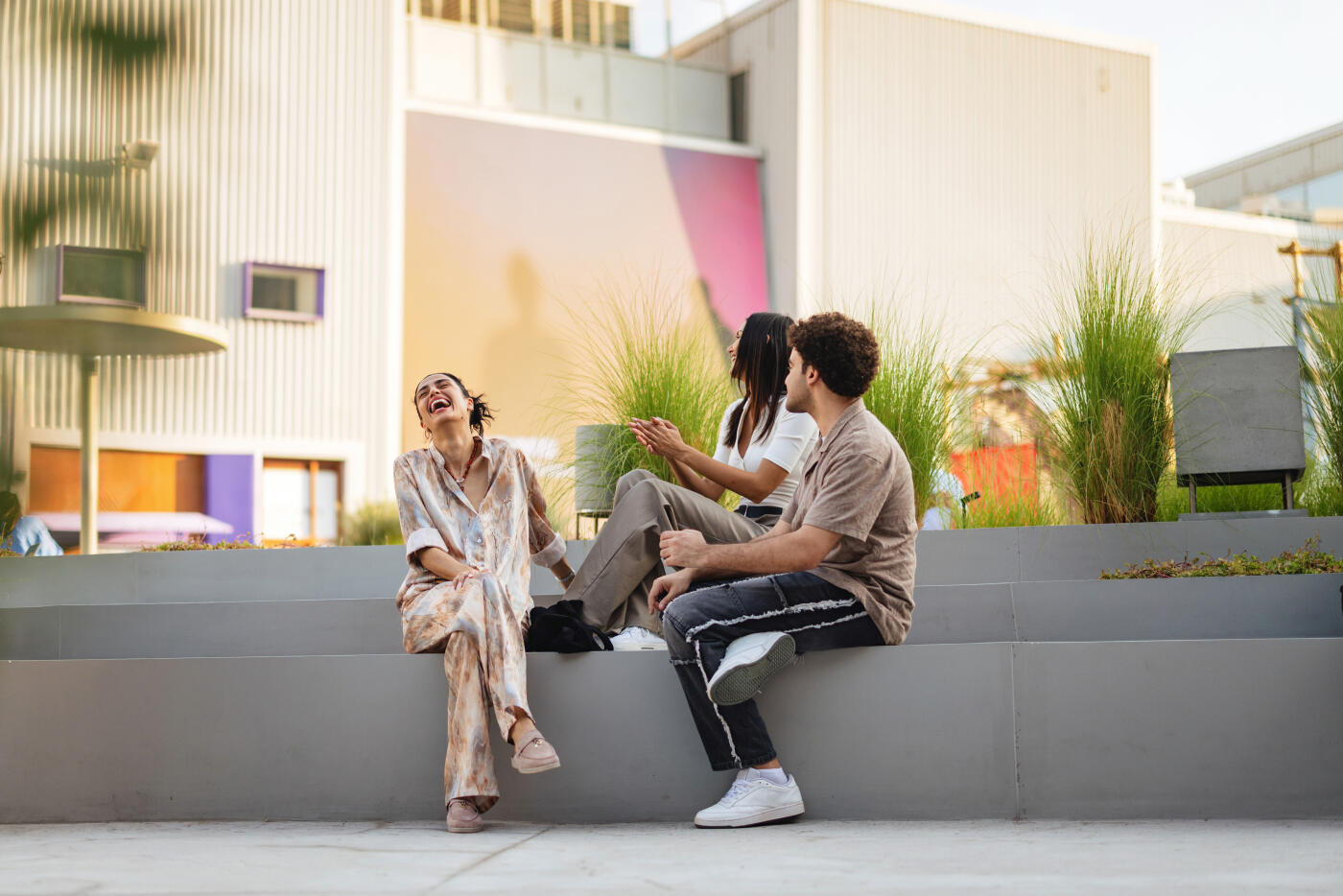 A diverse group of young adults enjoys a relaxed moment in a modern urban outdoor space in Dubai, dressed in casual and stylish clothing, surrounded by greenery and contemporary architecture under soft daylight.