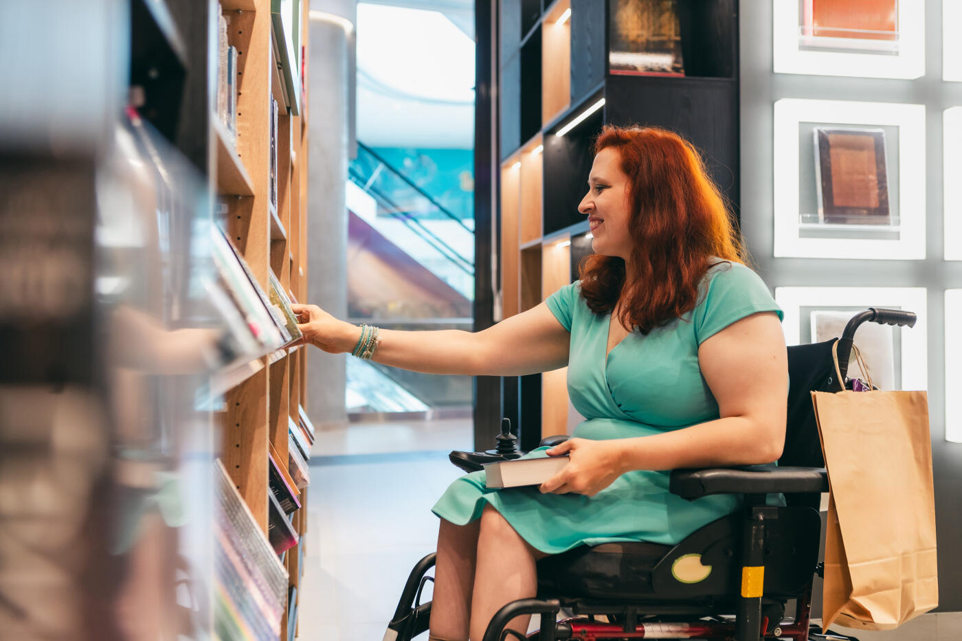 A happy woman with red hair sitting in an electric wheelchair reaching out to pick a book from a wooden bookshelf